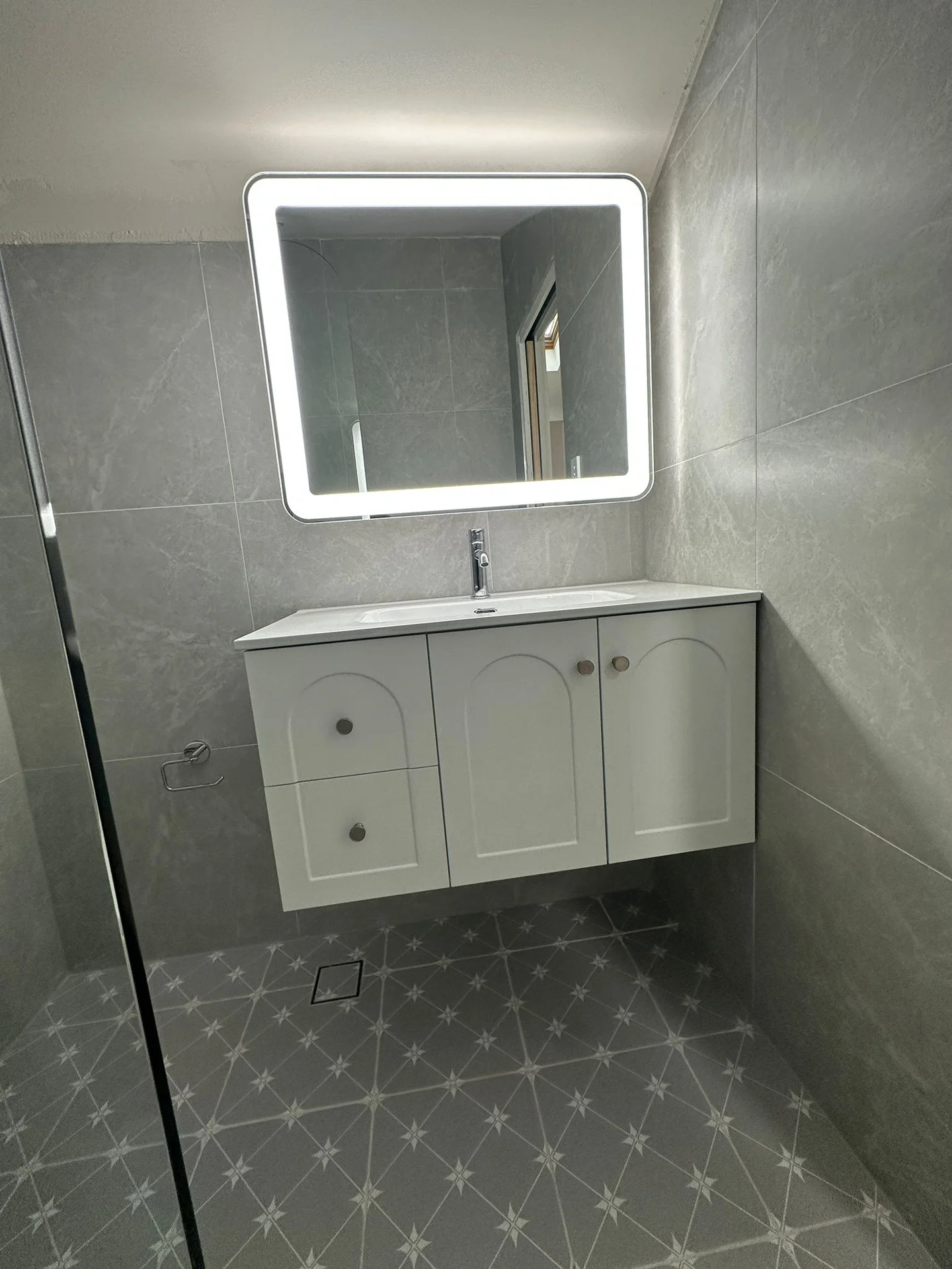 Grey bathroom wall tiles behind a white vanity and illuminated mirror
